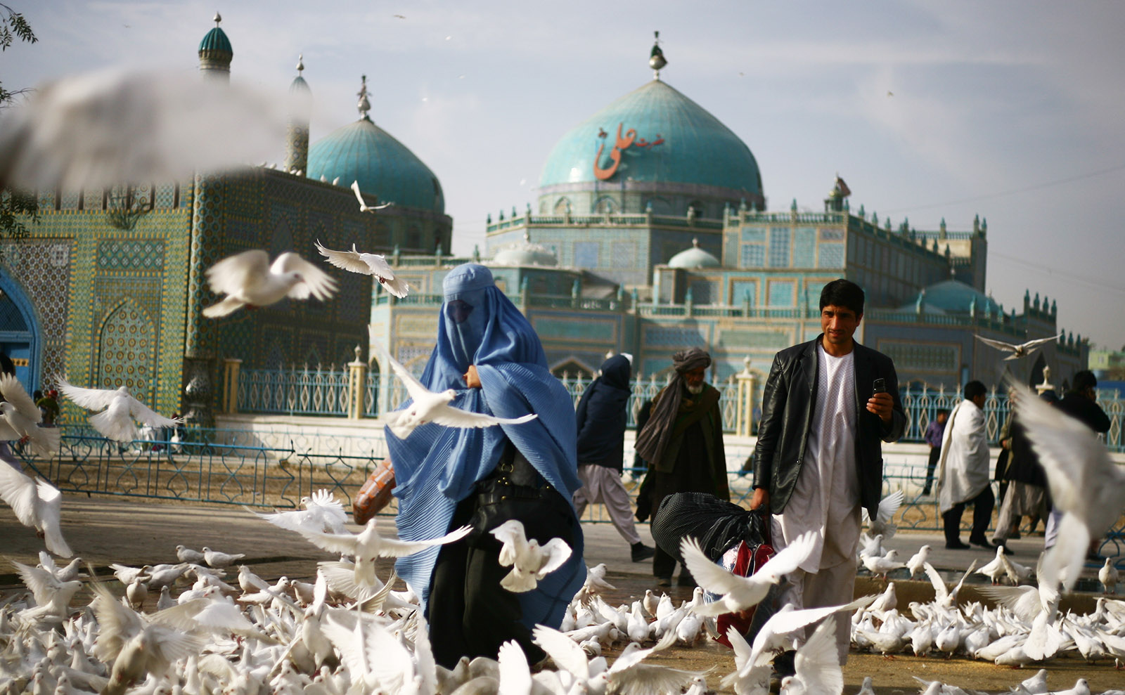 women wearing a blue veil in a square surrounded by white doves with a turquoise mosque in the background