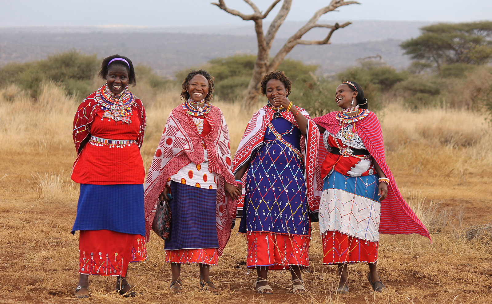 four kenyan women wearing traditional red and blue garments and smiling with the desert in the background