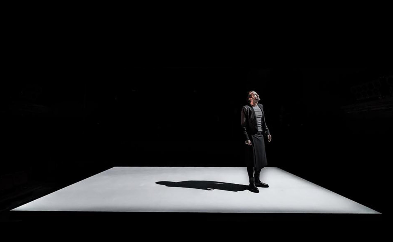 actor david tennant wearing gray and black standing on an empty stage in a spotlight