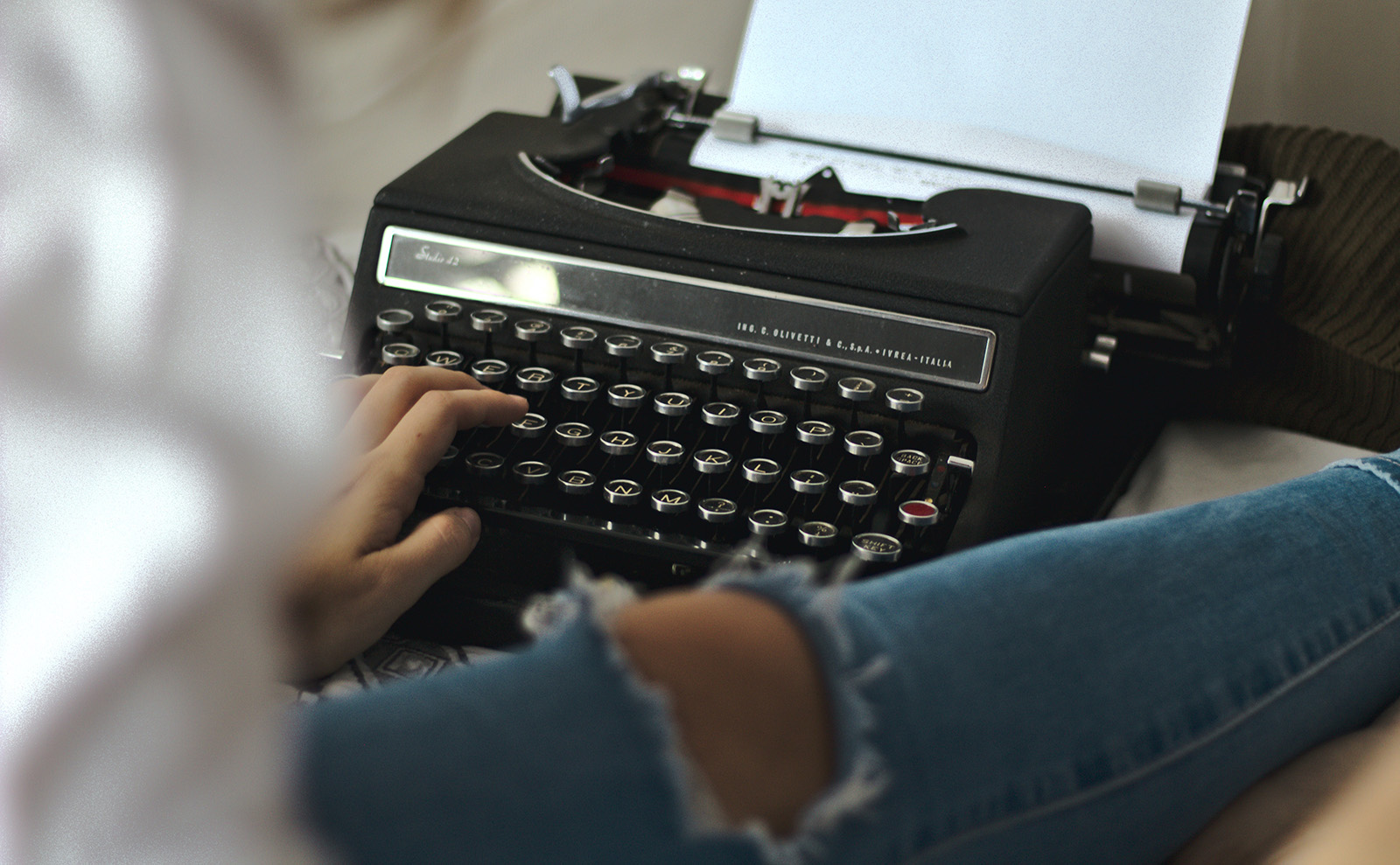  woman in a white shirt and torn blue jeans sitting on the floor in front of a typewriter